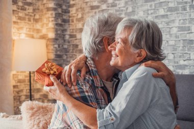 Evde birlikte eğlenen iki tatlı ve yaşlı yaşlının portresi. Olgun bir adam karısına yılbaşı ya da yıldönümü hediyesi veriyor. Şaşırmış emekli kadın kocasına sarılıyor.. 