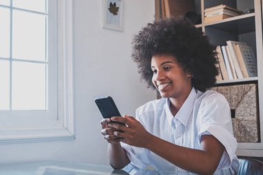 Kıvırcık saçlı, gülümseyen Afro-Amerikan kadın ofisinde oturuyor ve cep telefonuyla mesaj atıyor. İş yerinde cep telefonu kullanan mutlu bir iş kadını. Akıllı telefon kullanan kadın yönetici sosyal medya içeriğini tarıyor 