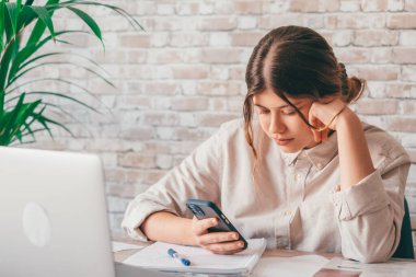 Bin yıllık kız öğrenciyi kapatın. Elinde telefonla oturmuş önemli bilgileri ya da verileri kontrol ediyor, kağıda not alıyor. Üniversite sınavına ya da sınava hazırlanan odaklanmış genç bir kız.. 