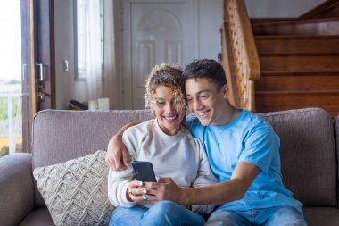 40 'lı yaşlarda gülümseyen anne yetişkin oğluyla birlikte akıllı telefon kullanıyor mutlu genç adam hafta sonunun keyfini çıkarıyor anneyle selfie çekiyor, evde eğleniyor. 