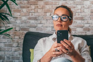 Modern lüks dairenin oturma odasındaki koltukta cep telefonu kullanarak gündelik kıyafetlerini giyen düşünceli genç bir kadın. Beyaz kadın internette sohbet ediyor ya da evde akıllı telefon kullanarak sosyal medya içeriğini izliyor. 