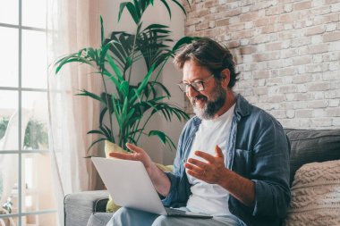 Mutlu olgun bir adam evdeki bilgisayarını kullanarak video izlerken koltukta dinleniyor. Beyaz erkek kanepede oturmuş internette sohbet ederken el kol hareketi yapıyor. Kendine güvenen bir adam sanal internet konferansı yapıyor. 