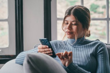 Genç ve güzel bir kadın evdeki kanepede oturmuş sohbet ediyor ve internette sörf yapıyor. İnternette akıllı telefonla eğlenen bir kadın. Sosyal medyada cep telefonuyla gülümseyen kız portresi 