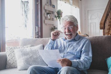 Heyecanlı son sınıf öğrencisi, elindeki belgelerden iyi haberleri okuyarak başarısını kutluyor. Yaşlı erkek zafer için tezahürat yapıyor. Kolları havada evin oturma odasındaki kanepede oturuyor.
