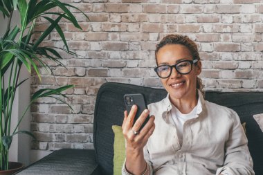 Evdeki rahat koltukta oturan gülümseyen bin yıllık beyaz kadın cep telefonu kullanıyor. Evinde rahat bir kanepede oturup internette sohbet eden mutlu genç bayan. Çevrimiçi konferansa katılan neşeli serbest yazar 