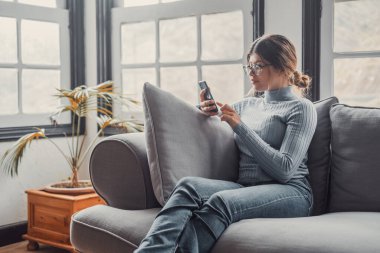 Genç ve güzel bir kadın evdeki kanepede oturmuş sohbet ediyor ve internette sörf yapıyor. İnternette akıllı telefonla eğlenen bir kadın. Sosyal medyada cep telefonuyla gülümseyen kız portresi 
