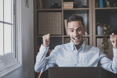 Şaşırmış Kafkasyalı genç işadamı ofisteki dizüstü bilgisayarla online olarak iyi haberler alarak başarıyı kutluyor. İş yerindeki başarıdan memnun olan erkek işletme yöneticisi