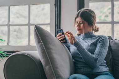 Genç ve güzel bir kadın evdeki kanepede oturmuş sohbet ediyor ve internette sörf yapıyor. İnternette akıllı telefonla eğlenen bir kadın. Sosyal medyada cep telefonuyla gülümseyen kız portresi 