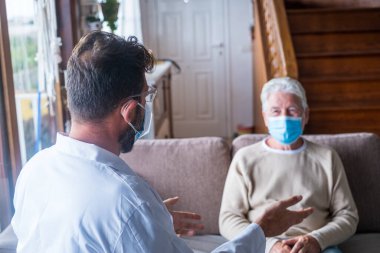 Erkek profesyonel doktor, tıbbi bakım ziyareti sırasında maske takarak kıdemli hastaya danışmanlık yapıyor. Genç bir doktor ve yaşlı bir ihtiyar kanepeye oturup tıbbi yardım sağlıyorlar. Yaşlı insanlar evde bakıyorlar. Covid ve Coronavirus pozitif test