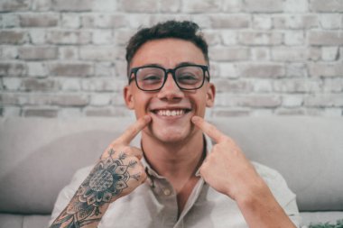 Genç, dövmeli bir adam gülümseyip kameraya bakıyor ve gülüşünü göstererek eğleniyor. Gözlük takan bir gencin portresi. 