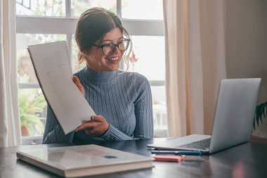 One young pretty woman studying and doing homework at home on the table. Female teenager using laptop or computer surfing the net indoor.   clipart