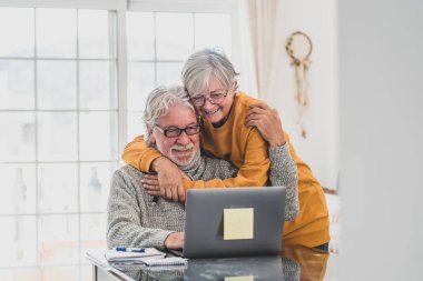 couple of two happy seniors working and using laptop together at home smiling and having fun together - lockdown lifestyle  clipart