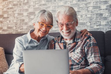 Kanepede oturup dizüstü bilgisayarla alışveriş yapan ve internette sörf yapan sevimli yaşlı insanlar. İki yetişkin insan oturma odasında gözlük takıyor ve teknolojinin tadını çıkarıyor. Yaşlıların birbirine gülen portresi.. 