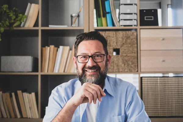 stock image Happy male trainer or teacher webinar speaker looking at camera giving online class lecture. Business executive or coach recording training video. Happy caucasian man sitting in front of bookshelf at office 