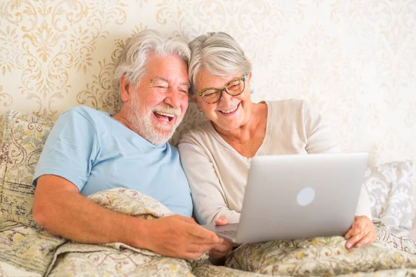 Old senior caucasian couple laughing and using laptop in the morning at bed in the bedroom at home. Elderly couple surfing and using social media on laptop at home.