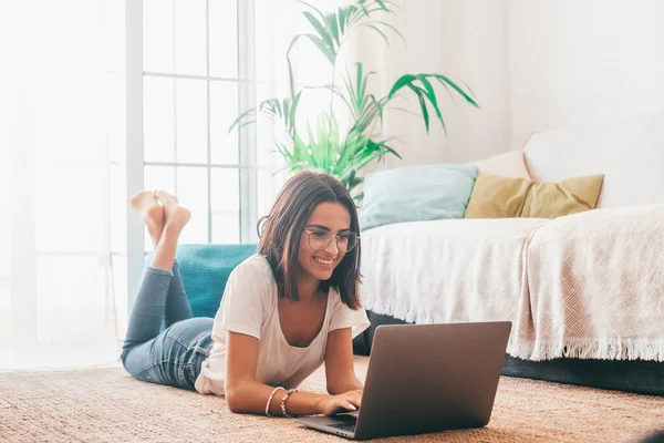 快乐的年轻女子戴着眼镜 躺在自家客厅的地毯上 一边用笔记本电脑打字 漂亮的女士用笔记本电脑消磨时光 在家里工作的自由职业者 — 图库照片