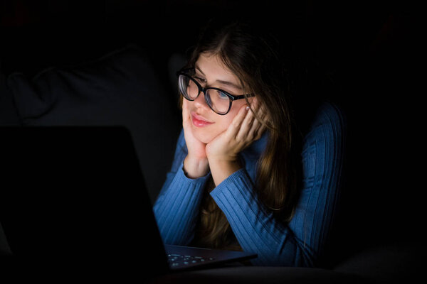 Одна счастливая жизнерадостная молодая женщина, пользующаяся ноутбуком поздно вечером в гостиной на диване дома. Онлайн-девушка в сети, пересматривает социальные сети. Улыбающийся подросток, использующий технологии и интернет. 