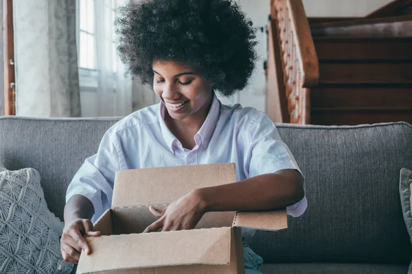 stock image African woman sitting on sofa in living room hold on lap big carton box unpacking parcel received from online shopping. Curious black lady unpacking cardboard parcel. Smiling female unboxing package 