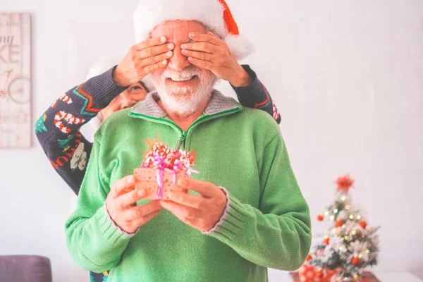 Senior couple in warm clothing and Santa hat, woman covering eyes of old husband with a surprise gift for him. Loving old romantic heterosexual couple celebrating Christmas festival together
