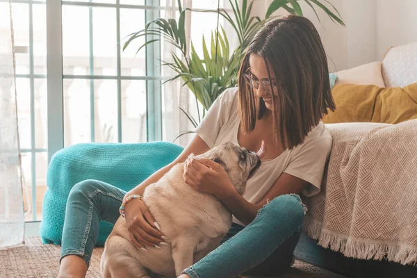 Portrait of beautiful woman in eyeglasses having fun with her pet pug dog sitting on floor in the living room of her house. Cheerful woman spending leisure time with her cute pet dog at home