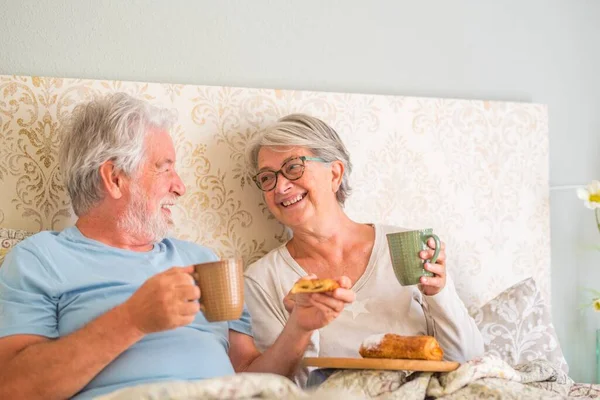 Old senior caucasian couple laughing and enjoying breakfast in the morning at bed in the bedroom at home. Elderly couple eating croissant and drinking coffee from cup for breakfast at home.