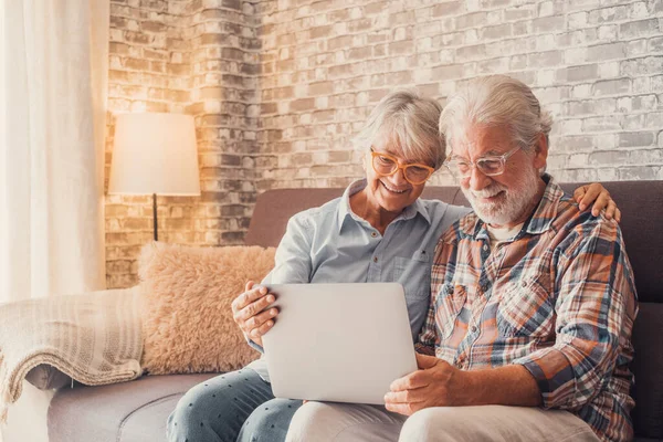 stock image Cute couple of old people sitting on the sofa using laptop together shopping and surfing the net. Two mature people wearing eyeglasses in the living room enjoying technology. Portrait of seniors laughing in love. 