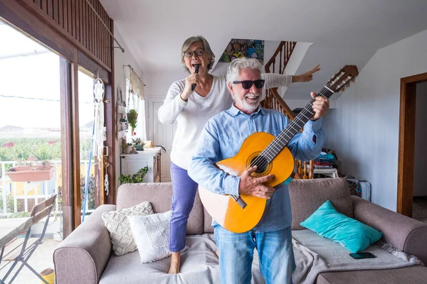 Happy and funny couple of old and mature people having fun and enjoying at home doing a party together singing and dancing playing the guitar indoor. Holiday or event celebrating concept.