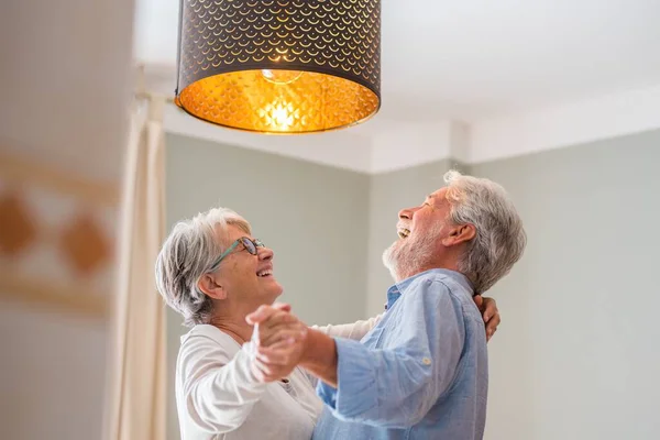 Cheerful senior couple dancing and laughing at home. Elderly happy couple celebrating by dancing in living room. Old couple having fun holding hands and enjoying dancing together at home