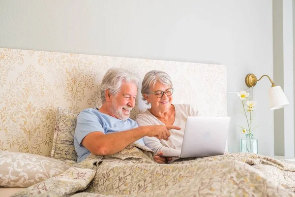 Old senior caucasian couple smiling and using laptop in the morning at bed in the bedroom at home. Elderly couple surfing and using social media on laptop at home. man pointing towards laptop screen