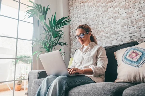 Young woman in casual clothes using laptop sitting on sofa in the living room of modern luxury apartment. Caucasian lady typing on keyboard chatting online or watching social media content at home