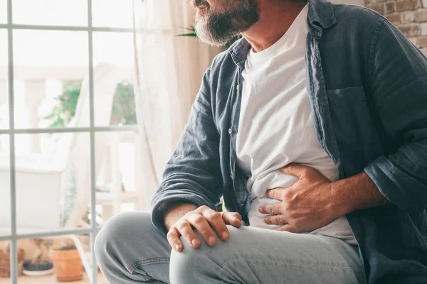 stock image Man suffering with severe stomach pain sitting at home. Hand of mature guy holding abdomen suffering from ache, diarrhea or indigestion problem. Caucasian guy pressing on belly on painful sensation 