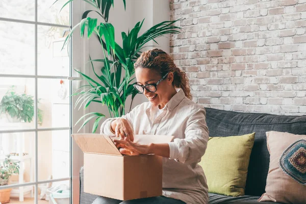 Woman unpacking delivery box opening package at home. Happy young lady looking at carton box while sitting on sofa in living room at modern apartment. Caucasian female checking out delivered stuff.