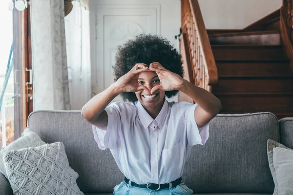 Happy african american woman sitting in living room connecting fingers of both hands making heart shape at home. Smiling black lady with curly hair making love heart sign sitting on comfortable sofa