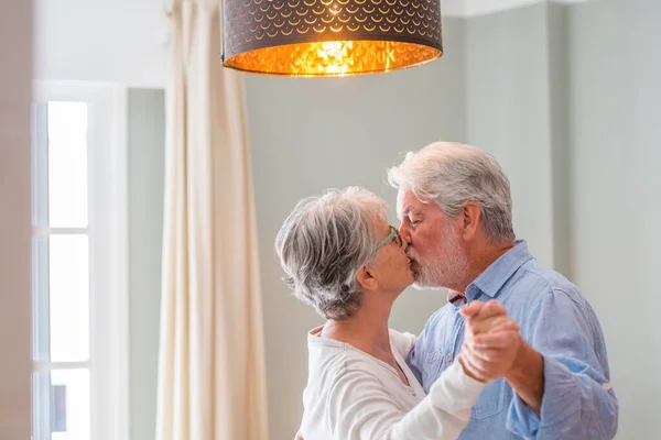 Cheerful senior couple kissing while dancing at home. Elderly happy couple dancing under chandelier hanging in living room. Romantic old couple kissing each other at home