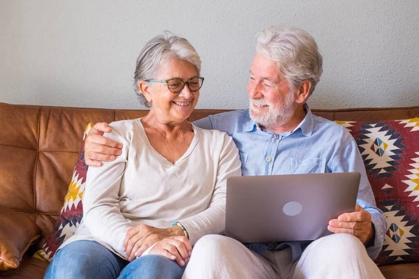 Cheerful senior couple using laptop while sitting on sofa and smiling. Elderly happy couple relaxing while surfing on laptop sitting in living room. Old couple watching media content using laptop