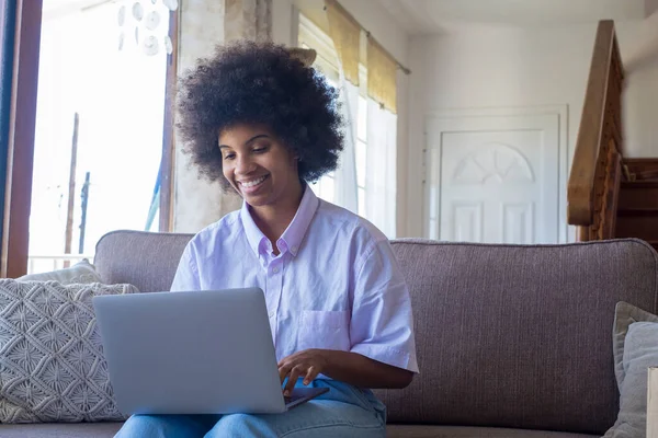 Smiling millennial african american woman using laptop sitting on comfortable sofa at home. Pretty happy black lady working from home on laptop sitting on couch in living room.