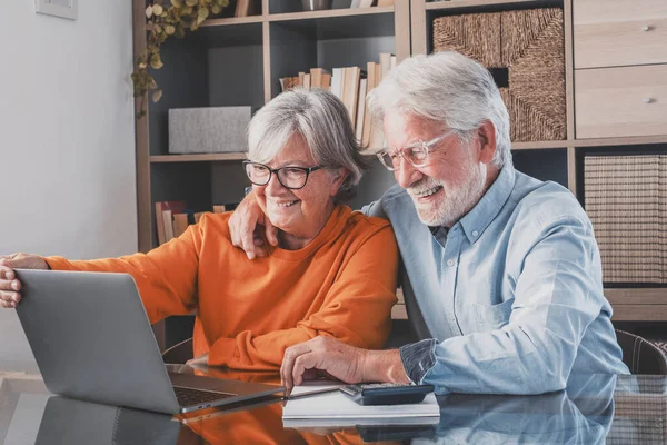 Elderly man and woman having fun using laptop at office. Smiling old husband and wife doing home finance online. Senior couple watching media content and enjoying at home