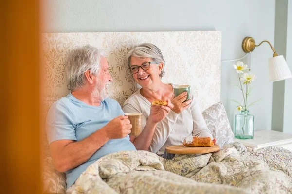 Old senior caucasian couple laughing and enjoying breakfast in the morning at bed in the bedroom at home. Elderly couple eating croissant and drinking coffee from cup for breakfast at home