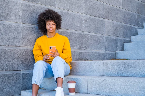 Beautiful african american woman in yellow t-shirt and curly hair using mobile phone outdoors on urban city staircase. Black lady text messaging on smartphone while sitting on steps with disposable coffee cup