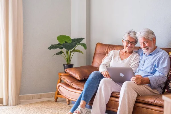 Cheerful senior couple using laptop while sitting on sofa and smiling. Elderly happy couple relaxing while surfing on laptop sitting in living room. Old couple watching media content using laptop