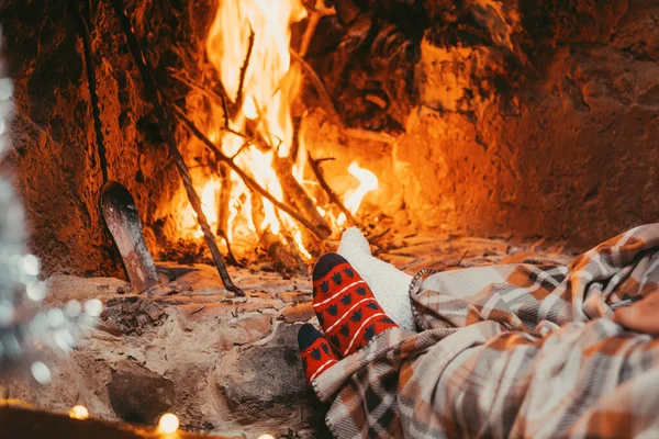 Low section legs of husband and wife covered in socks and warm blanket relaxing in front of burning fireplace during winter christmas holiday. Pair of feet in woolen socks by the Christmas fireplace