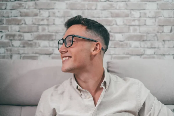 Portrait of one young attractive man looking at the window smiling and having fun sitting on the sofa. Happy male teenager wearing eyeglasses enjoying.