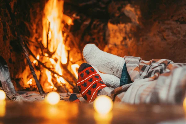 Low section legs of husband and wife covered in socks and warm blanket relaxing in front of burning fireplace during winter Christmas holiday. Pair of feet in woolen socks by the Christmas fireplace