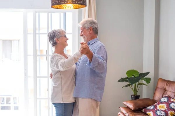 Romantic loving senior couple holding hands enjoying dancing together in the living room of house, Elderly happy couple celebrating by dancing in living room.