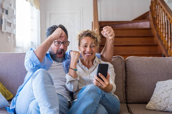 Astonished caucasian couple celebrating online success using laptop sitting on sofa at home. Joyful wife and husband surprised by good unbelievable news, huge lottery jackpot win