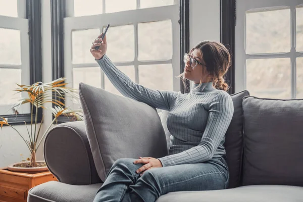 Young beautiful woman sitting on the sofa at home chatting and surfing the net. Female person having fun with smartphone online. Portrait of girl smiling taking a selfie to post it on the social media.