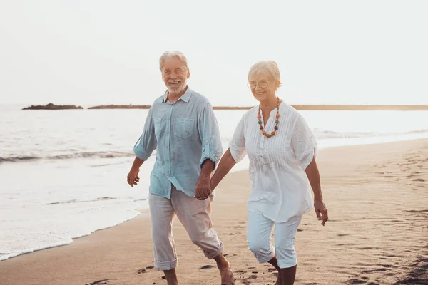 Couple of old mature people walking on the sand together and having fun on the sand of the beach enjoying and living the moment. Two cute seniors in love having fun. Barefoot walking on the water