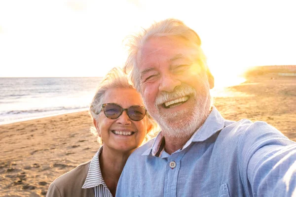 stock image Portrait of couple of mature and old people enjoying summer at the beach looking to the camera taking a selfie together with the sunset at the background. Two active seniors traveling outdoors. 