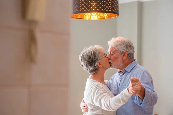 Romantic loving senior couple holding hands while kissing and enjoying dancing together in the living room of house, Elderly happy couple celebrating by doing dance at home. Old husband and wife having fun time at modern apartment