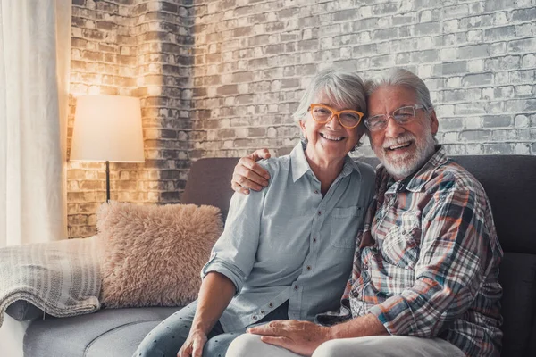 Cute and beautiful couple of old people smiling and looking at the camera having fun at home together. Portrait of seniors wearing eyeglasses sitting on sofa enjoying and relaxing.